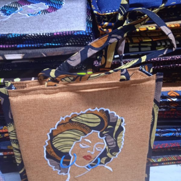 A beautiful celebration of heritage and style. This tan-colored bag features a stunning embroidered portrait of an African woman adorned in a vibrant headwrap and bold blue hoop earrings. Striking Artistry: A bold, artistic embroidered portrait that serves as a conversation starter. Natural Texture: Made with a burlap-textured fabric that offers a rustic yet sophisticated look. Coordinated Design: The patterned straps and side trim perfectly complement the colors in the central portrait. Unique Detail: Every bag features unique pattern variations on the handles, making each piece one-of-a-kind.